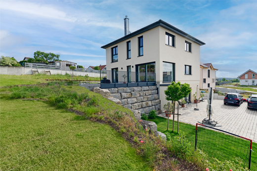 Freiraum-Haus von Aischtaler Holzhaus in Hanglage mit heller Fassade, großer Terrasse, Natursteinmauer und Blick auf umliegende Bebauung.