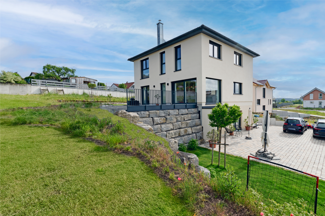 Freiraum-Haus von Aischtaler Holzhaus in Hanglage mit heller Fassade, großer Terrasse, Natursteinmauer und Blick auf umliegende Bebauung.