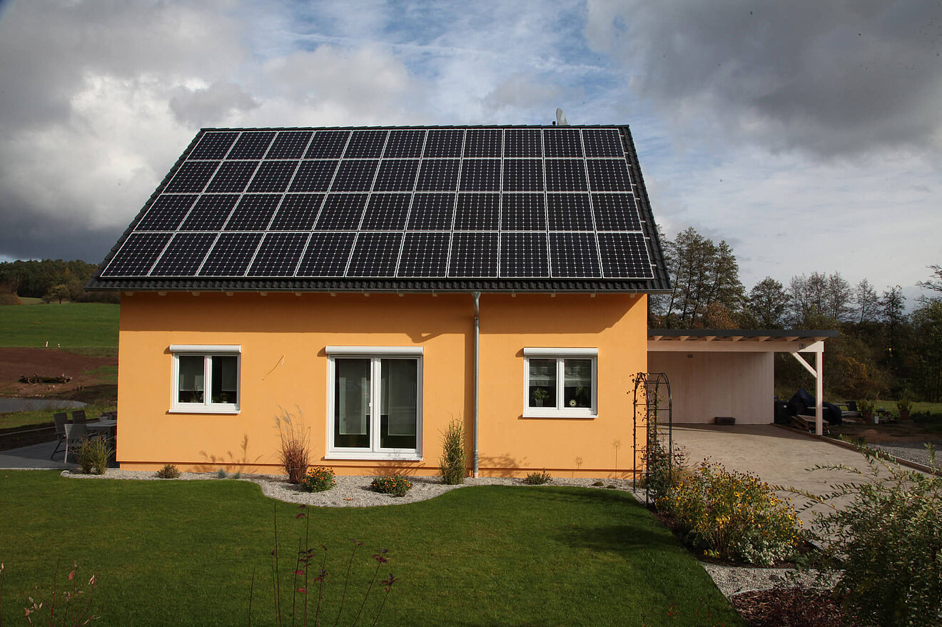 Aischtaler Holzhaus Sonnenenergiehaus mit großer Photovoltaikanlage auf dem Satteldach und sonnengelber Fassade, seitlicher Carport und gepflegter Garten