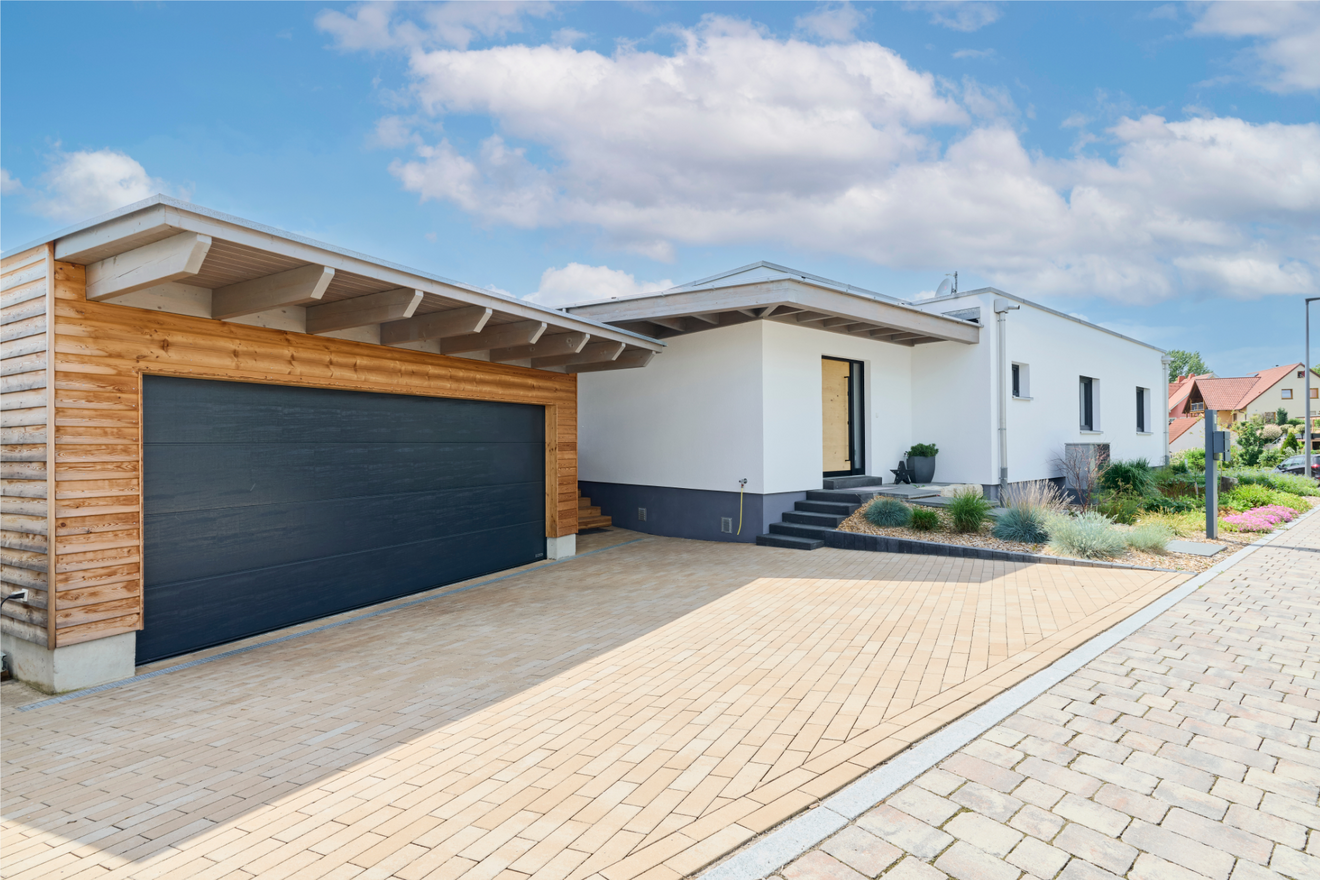 Modernes Kubikhaus von Aischtaler Holzhaus mit weißer Fassade, dunkler Eingangstür, vorgelagerter Holzgarage und gepflasterter Einfahrt.