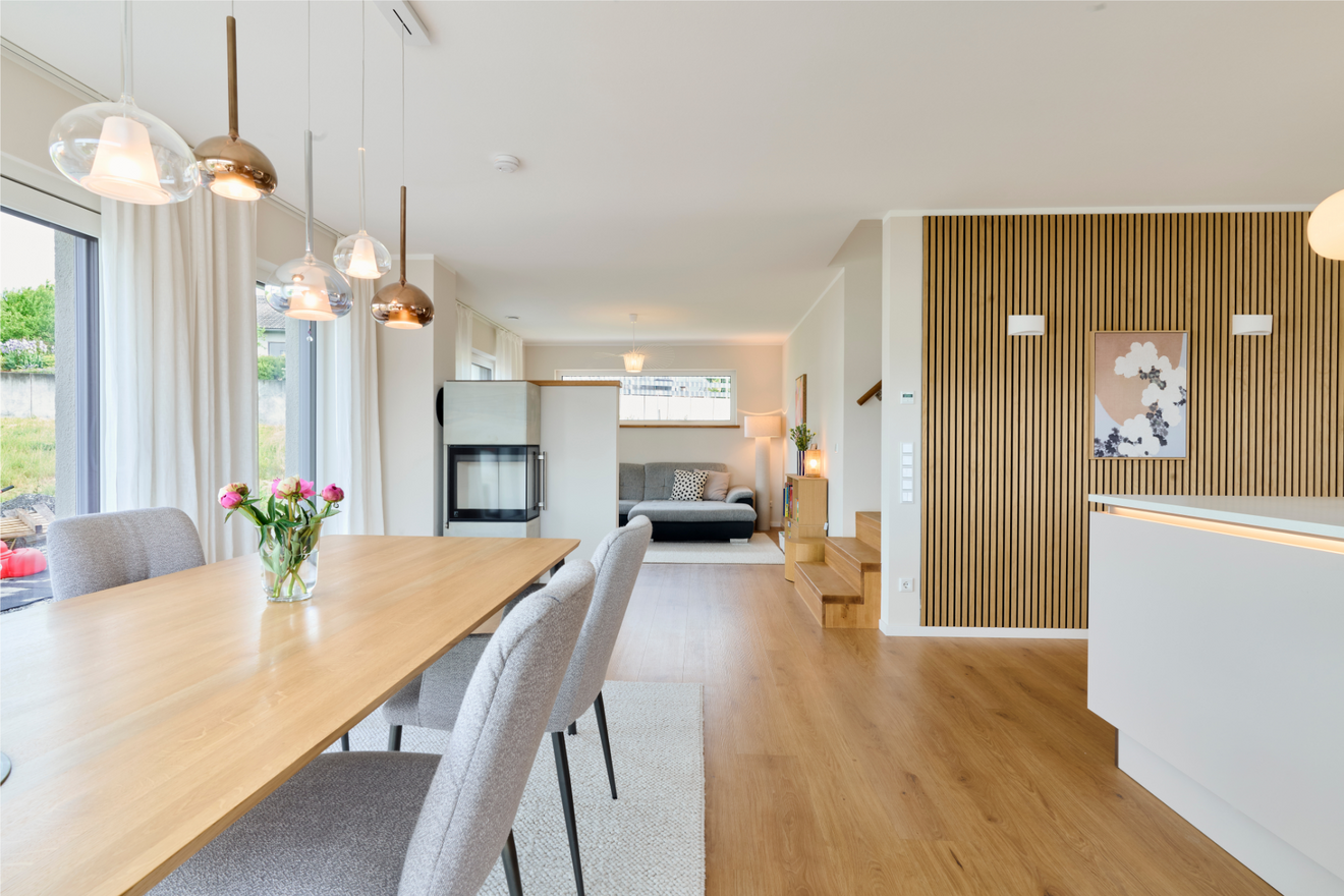 Heller Raum im Freiraum-Haus von Aischtaler Holzhaus mit Esstisch, Pendelleuchten, Kamin, Holzfußboden und Wandverkleidung aus vertikalen Holzleisten.