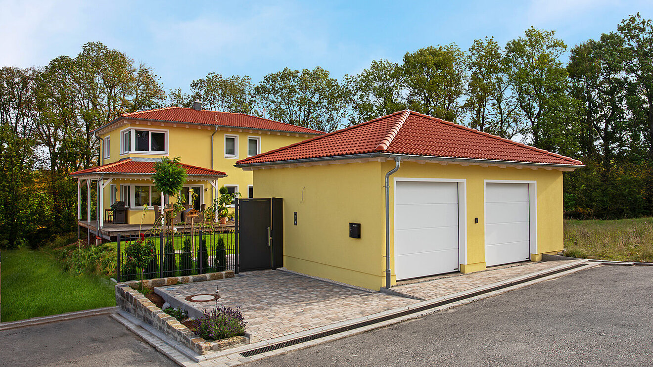 Toskanahaus von Aischtaler Holzhaus – gelbe Fassade mit rotem Walmdach, Terrasse und Doppelgarage im Grünen