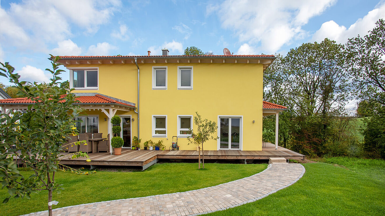 Toskanahaus von Aischtaler Holzhaus – gelbe Fassade mit rotem Walmdach, große Terrasse und Gartenansicht