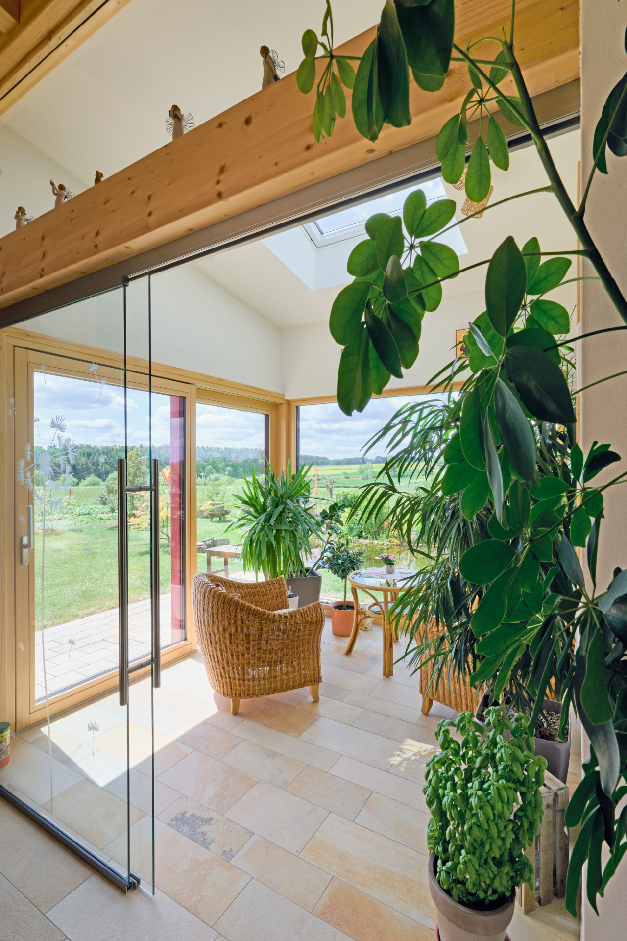 Sonnenlichter Wintergarten im Naturnah-Haus von Aischtaler Holzhaus mit vielen Zimmerpflanzen, Rattansesseln und Blick auf eine grüne Landschaft.