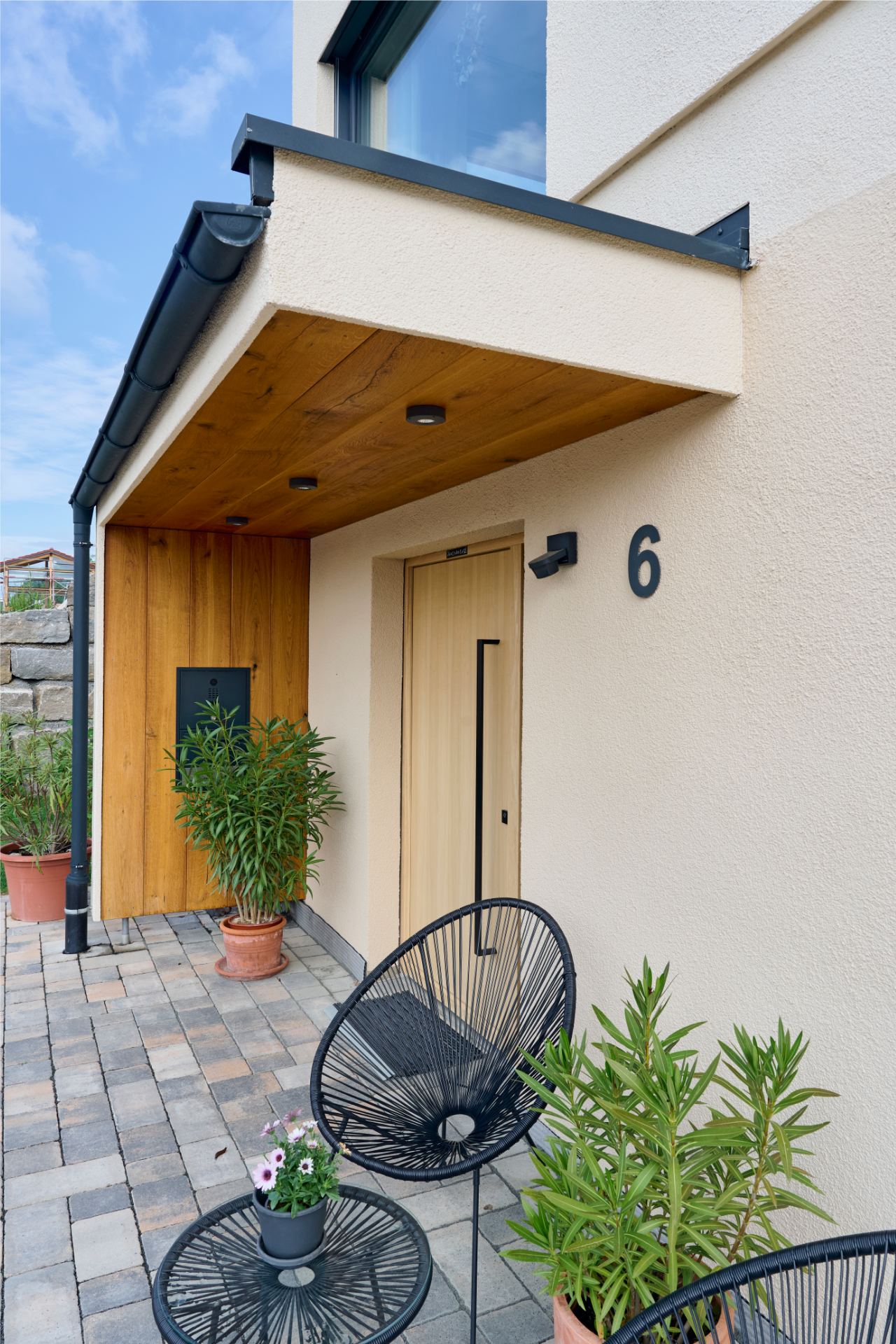 Eingangsbereich eines Freiraum-Hauses von Aischtaler Holzhaus mit heller Putzfassade, Holzdecke, moderner Haustür, Topfpflanzen und schwarzem Stuhlset.