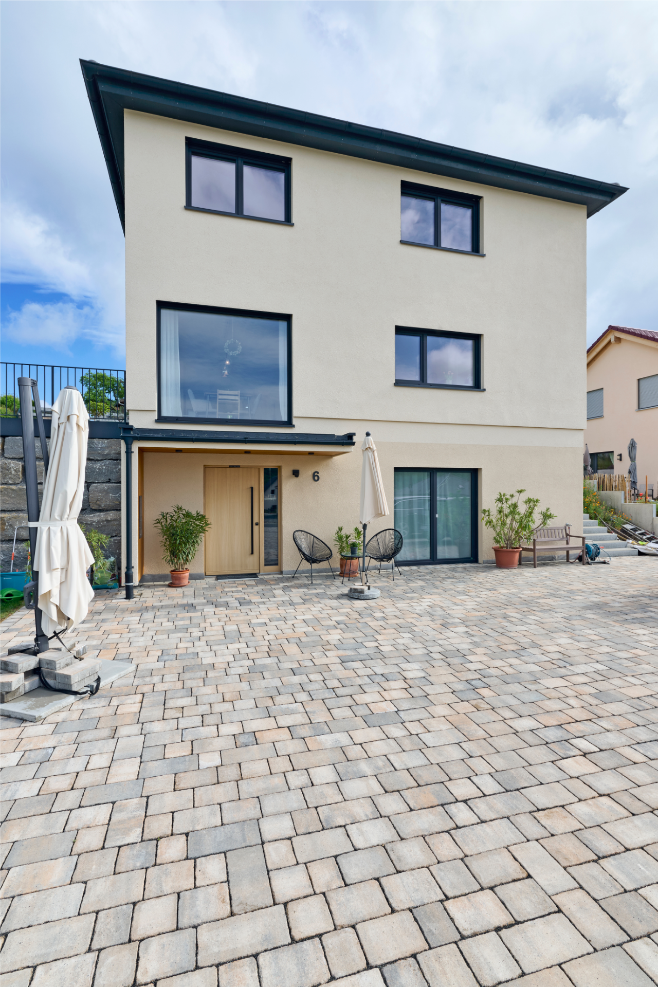Rückseite eines Freiraum-Hauses von Aischtaler Holzhaus mit heller Fassade, großem Fenster, Terrasse mit Pflasterung und möbliertem Eingangsbereich.