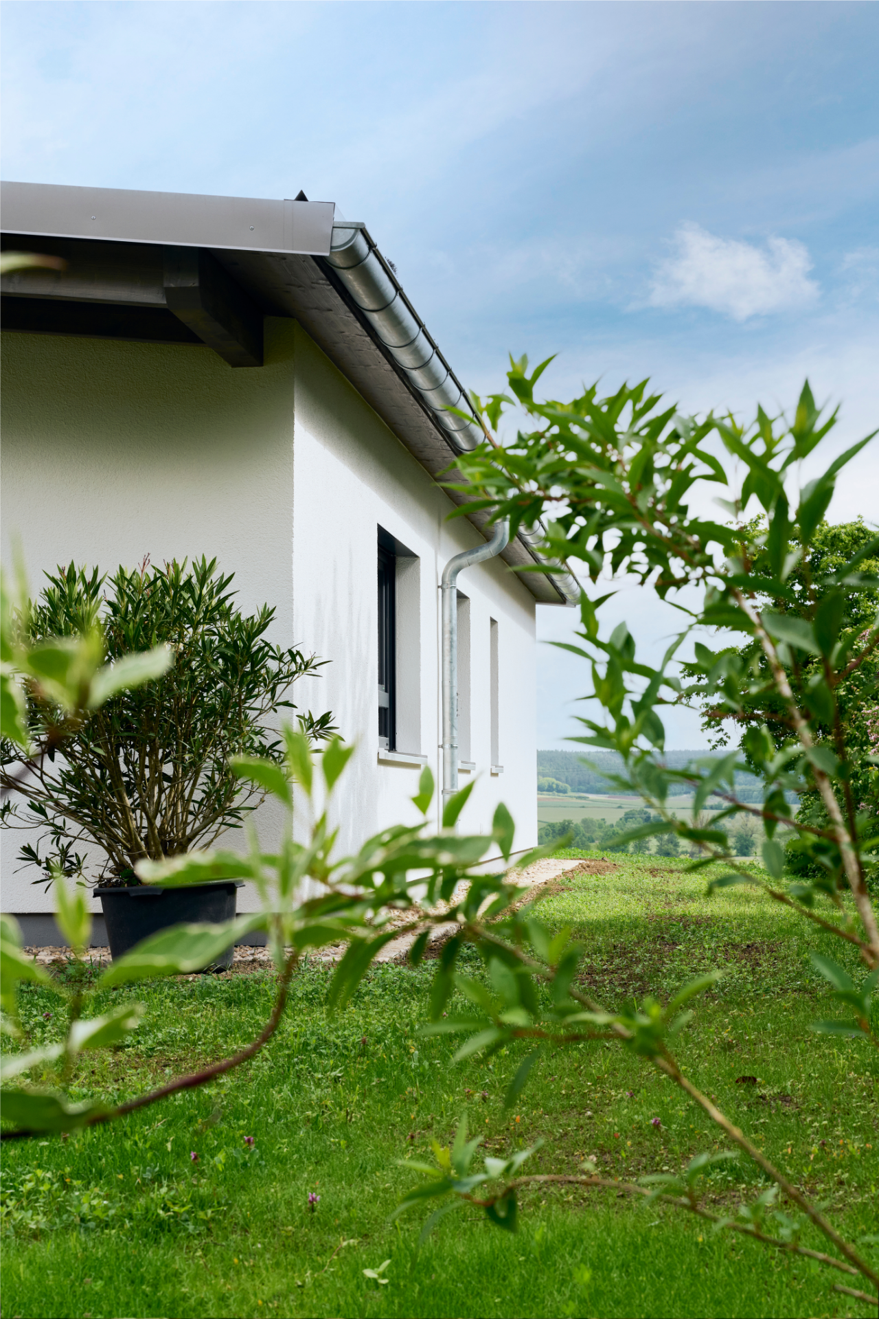 Weiße Außenwand des Tandemhauses von Aischtaler Holzhaus mit schmalen Fenstern und Garten, dahinter Felder und Hügel.
